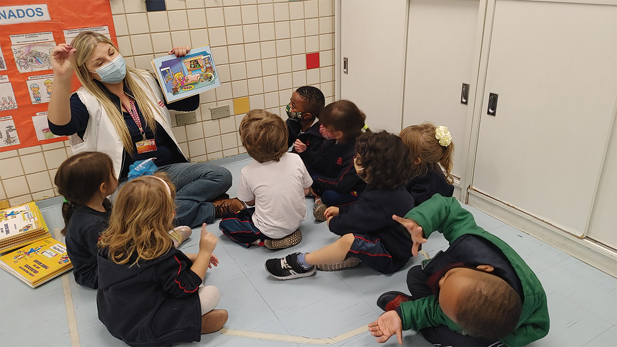 Foto de alunos sentados observando ilustração de livro que professora está mostrando