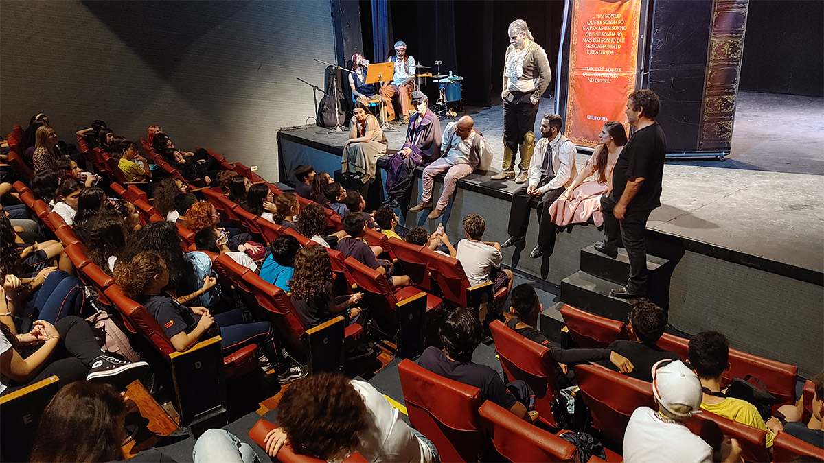 Foto de alunos reunidos em anfiteatro. Pessoas no palco conversam com eles